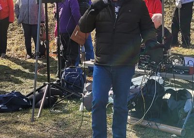 man holding microphone in front of outdoor pavilion tent with audio equipment behind him and onlookers behind