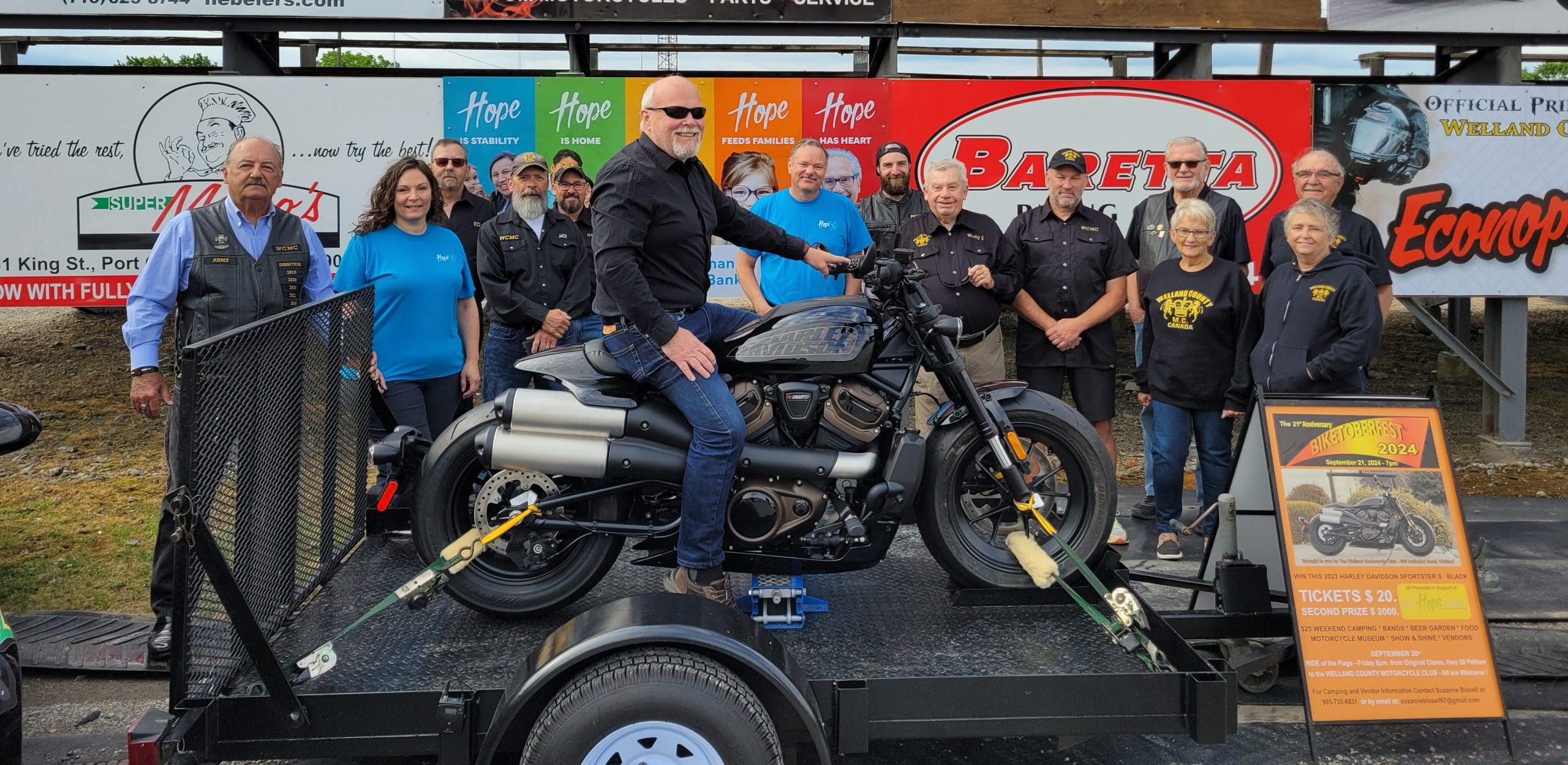 Group photo with the Welland County Motorcycle Club members, representatives from The Hope Centre and Mayor Campion sitting on a motorcycle that is being raffled for Biketoberfest.